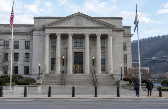 A wide shot shows the stone facade of a classical courthouse building with two flagpoles and two people walking toward the entrance.