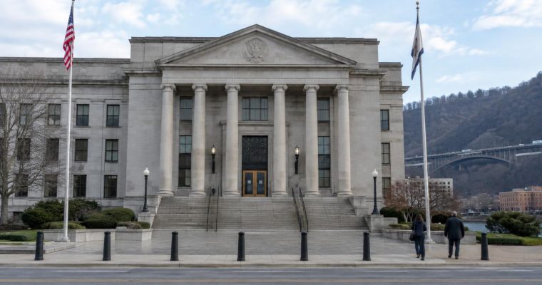 A wide shot shows the stone facade of a classical courthouse building with two flagpoles and two people walking toward the entrance.